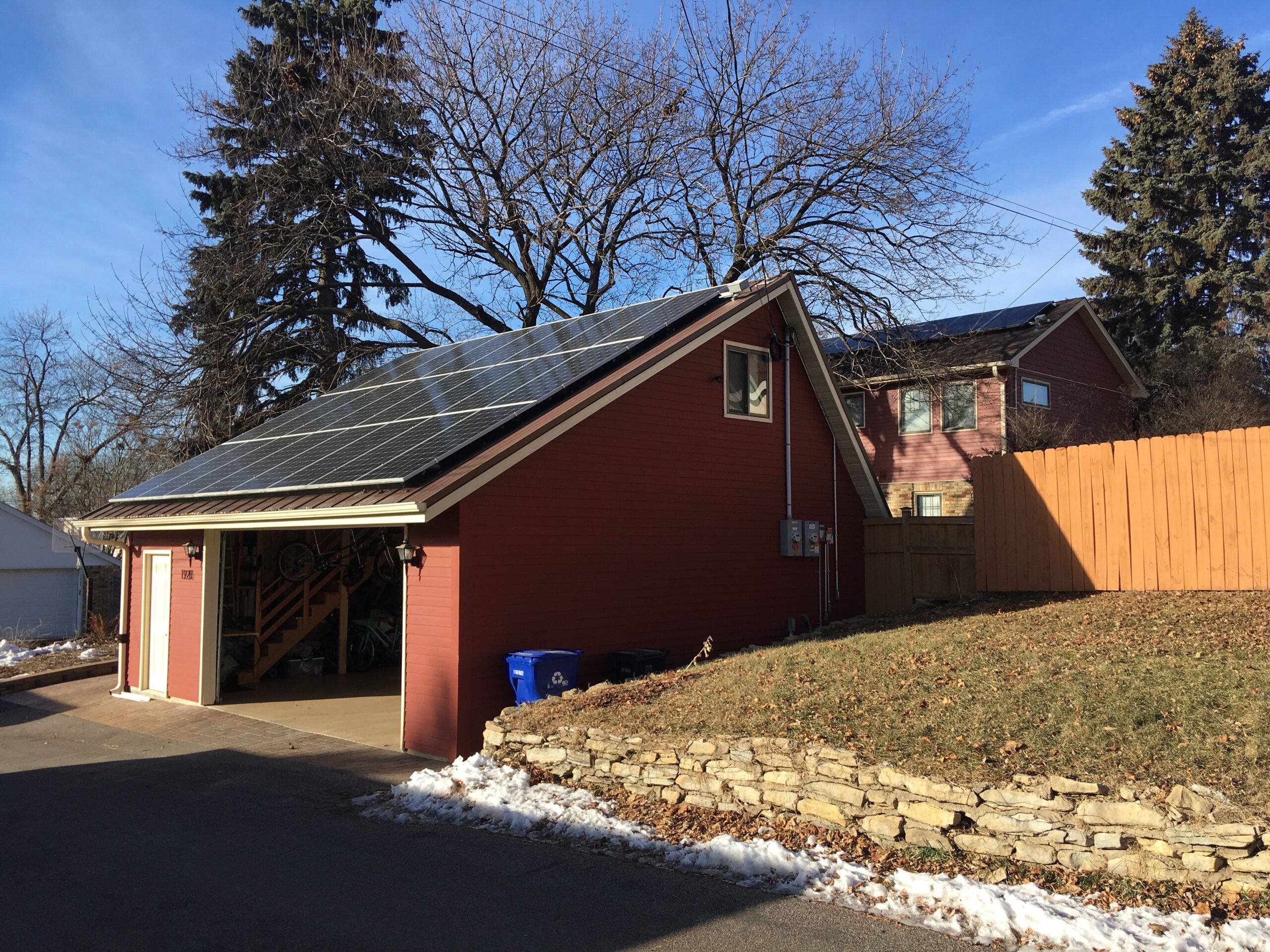 A red garage with the door open. Mid-winter in Minnesota with only a little bit of snow on the ground. There are solar panels on the roof of the garage, and it is very sunny out. There is a low fence acting as a retaining wall for part of the yard, with grass showing. In the background is a tall wooden fence and a single-family home.